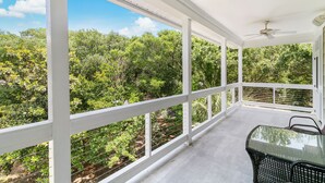Un balcón cubierto con mesa y sillas de vidrio, barandillas blancas y vistas a abundante vegetación.