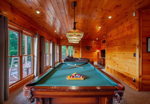 A pool table with a cue ball and billiard balls, surrounded by wooden walls and large windows.