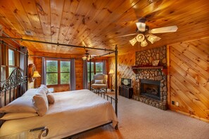 A log cabin bedroom with a four-poster bed, a stone fireplace, and a view of the outdoors through large windows. - 5 King Suite Cabin – Mountain Views, Creek Access & Hot Tub (Blue Ridge)