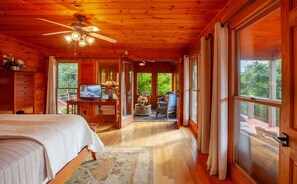 L’intérieur d’un chalet en bois comprend un lit, un téléviseur et un balcon offrant une vue sur la verdure.