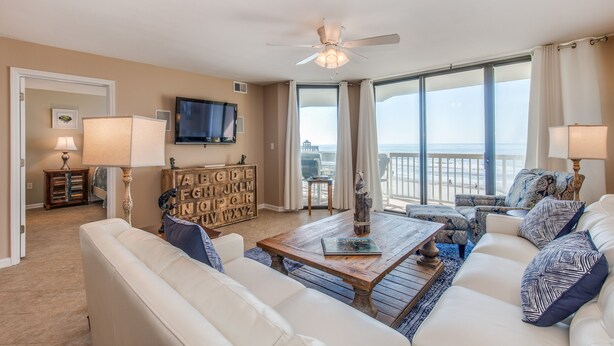 A spacious living room with a large window offering a view of the ocean, a ceiling fan, and a flat-screen TV mounted on the wall. - Stay @ Ocean's Nine on Folly Beach (Folly Beach)