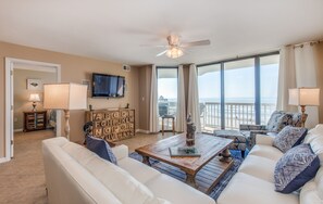 A spacious living room with a large window offering a view of the ocean, a ceiling fan, and a flat-screen TV mounted on the wall.