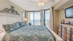 A bedroom with a large bed, a ceiling fan, a TV, and a view of the ocean. - Stay @ Ocean's Nine on Folly Beach (Folly Beach)