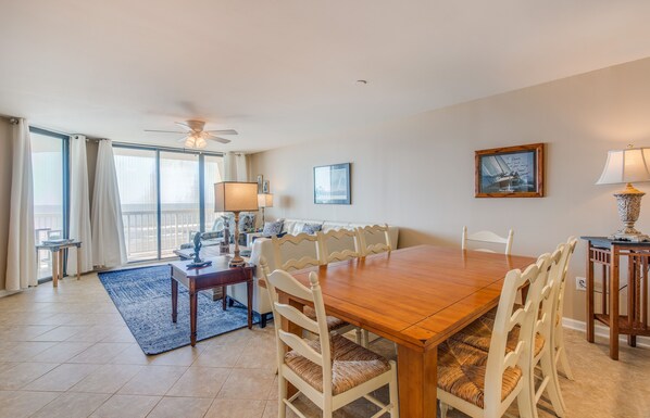 A dining area with a wooden table, white chairs, a ceiling fan, and a view through large windows.