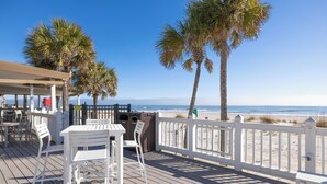 A wooden deck with white tables and chairs, overlooking a beach with palm trees and a clear blue sky. - Stay @ Serenity by the Sea on Isle of Palms (Isle of Palms)