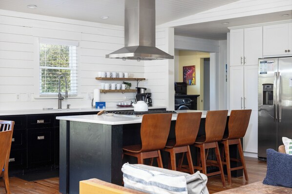 A modern kitchen with a central island, bar stools, a stainless steel refrigerator, and a window with blinds.