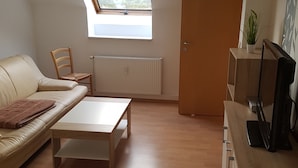 Een ruimtelijk ingerichte zithoek met een bank, een bijzettafeltje, een radiator, een deur en een televisie op een houten standaard.