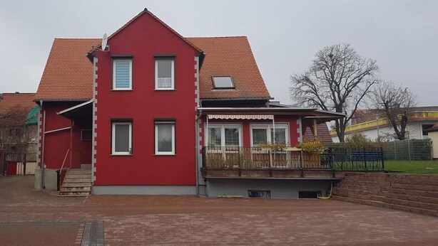 Een rode, twee verdiepingen tellende woning met een schuin dak, een klein portaaltje en een omheinde tuin.