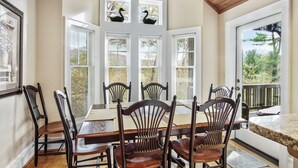 Un comedor con mesa y sillas de madera, ventanas grandes con contraventanas y vistas a la vegetación.