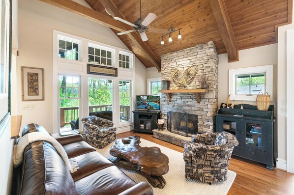 A cozy living room with a stone fireplace, leather sofa, patterned chairs, and a television.