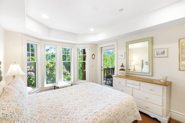 A bedroom with a large bed, a dresser, a mirror, and a window with a view of greenery.