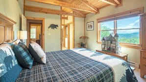 Une chambre avec un grand lit, des poutres apparentes en bois au plafond, une décoration murale représentant un ours, un fauteuil à bascule et une fenêtre donnant sur l’extérieur.