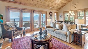 A spacious living room with a high ceiling, wooden beams, a large window, and a cozy seating area with a couch and armchairs. - Grand Oaks RS6- Mountain Condo, Gated Resort, Indoor/Outdoor Pools & Hot Tub (Banner Elk)