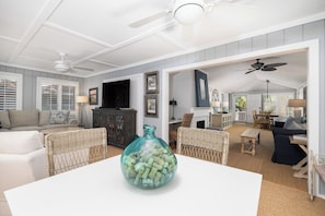 A spacious living room with a white ceiling fan, a large glass vase on a white table, and a dining area with wicker furniture.