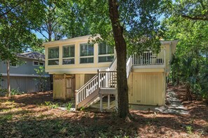 A two-story house with a staircase leading to a balcony, surrounded by trees and a wooden fence.