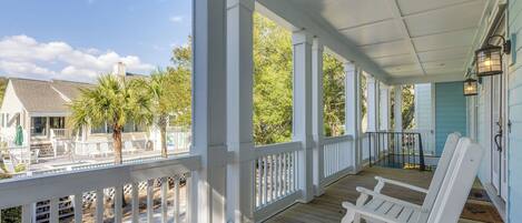 A covered porch with white railings, wooden flooring, and white Adirondack chairs.
