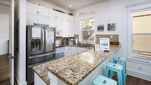 Une cuisine moderne avec des meubles blancs, des comptoirs en granit, un réfrigérateur en acier inoxydable et un four encastré. On y trouve deux tabourets de bar turquoise et une fenêtre avec des stores.