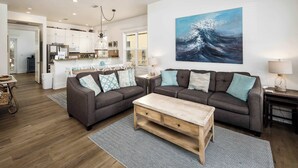Un salon spacieux avec un grand canapé d’angle gris, une table basse en bois et une cuisine en arrière-plan.