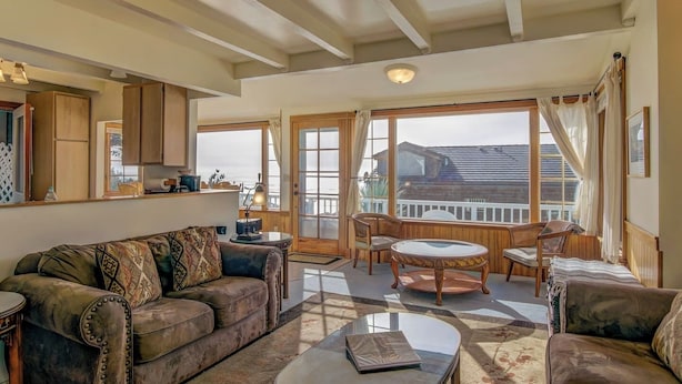 A spacious living room with a sofa, a coffee table, and a dining area with chairs. There is a large window providing a view of the outside.