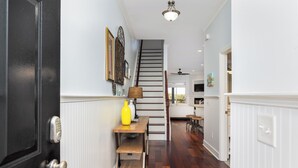 A hallway with a wooden floor, a staircase, a door with a keypad, and a table with a lamp and a vase.