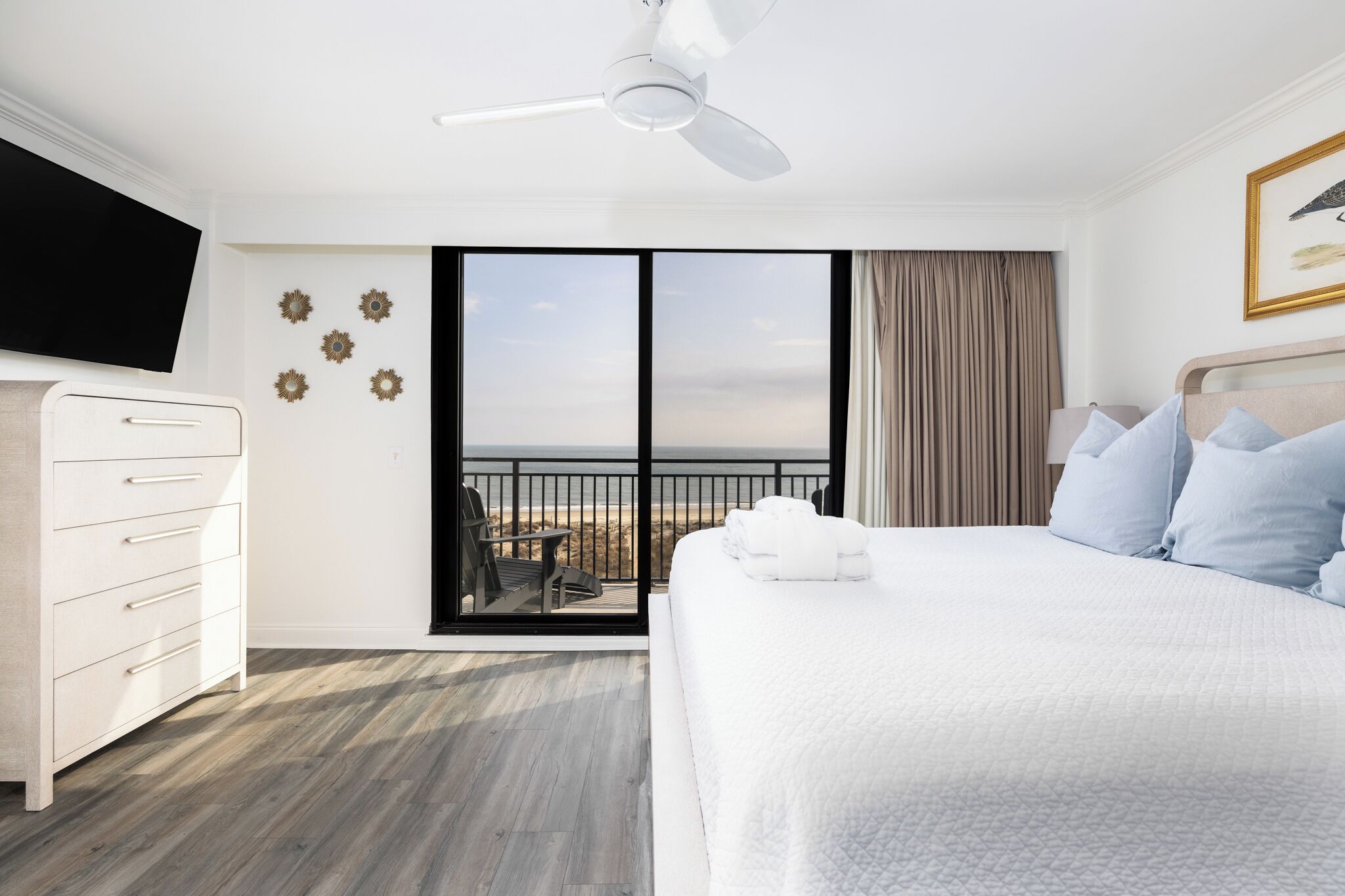 A hotel room with a bed, a dresser, a ceiling fan, a TV, and a balcony with a view of the beach.