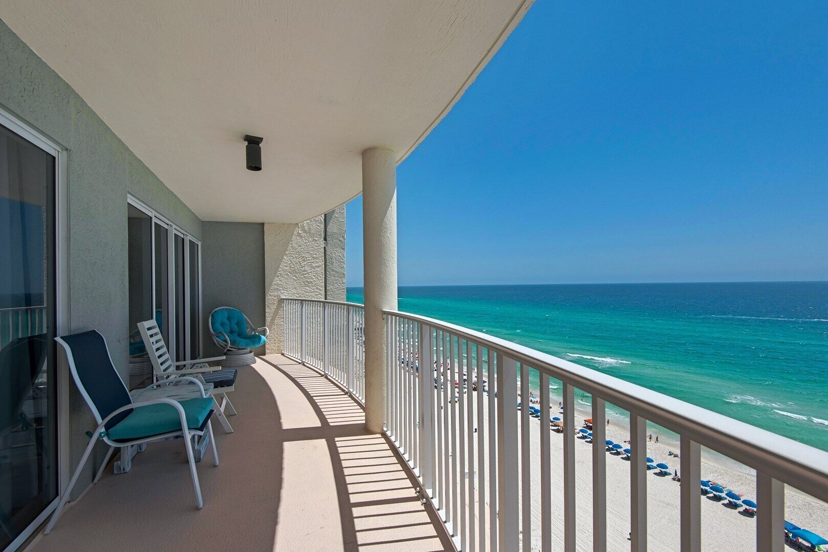 A balcony with two chairs and a table, overlooking a beach with umbrellas and people.