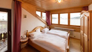 Ein schlicht eingerichtetes Schlafzimmer mit Holzbettrahmen, zwei Betten, einem Fenster mit Vorhängen, einem Heizkörper und einem Holzschrank.
