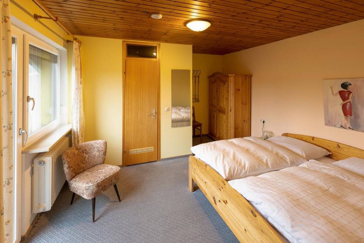 A bedroom with two beds, a chair, a radiator, a wooden wardrobe, and a painting on the wall.