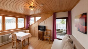 Ein gemütliches Zimmer mit Holzbalkendecke, einem Esstisch mit Stühlen, einer Couch, einem Fernseher und Seeblick durch große Fenster.