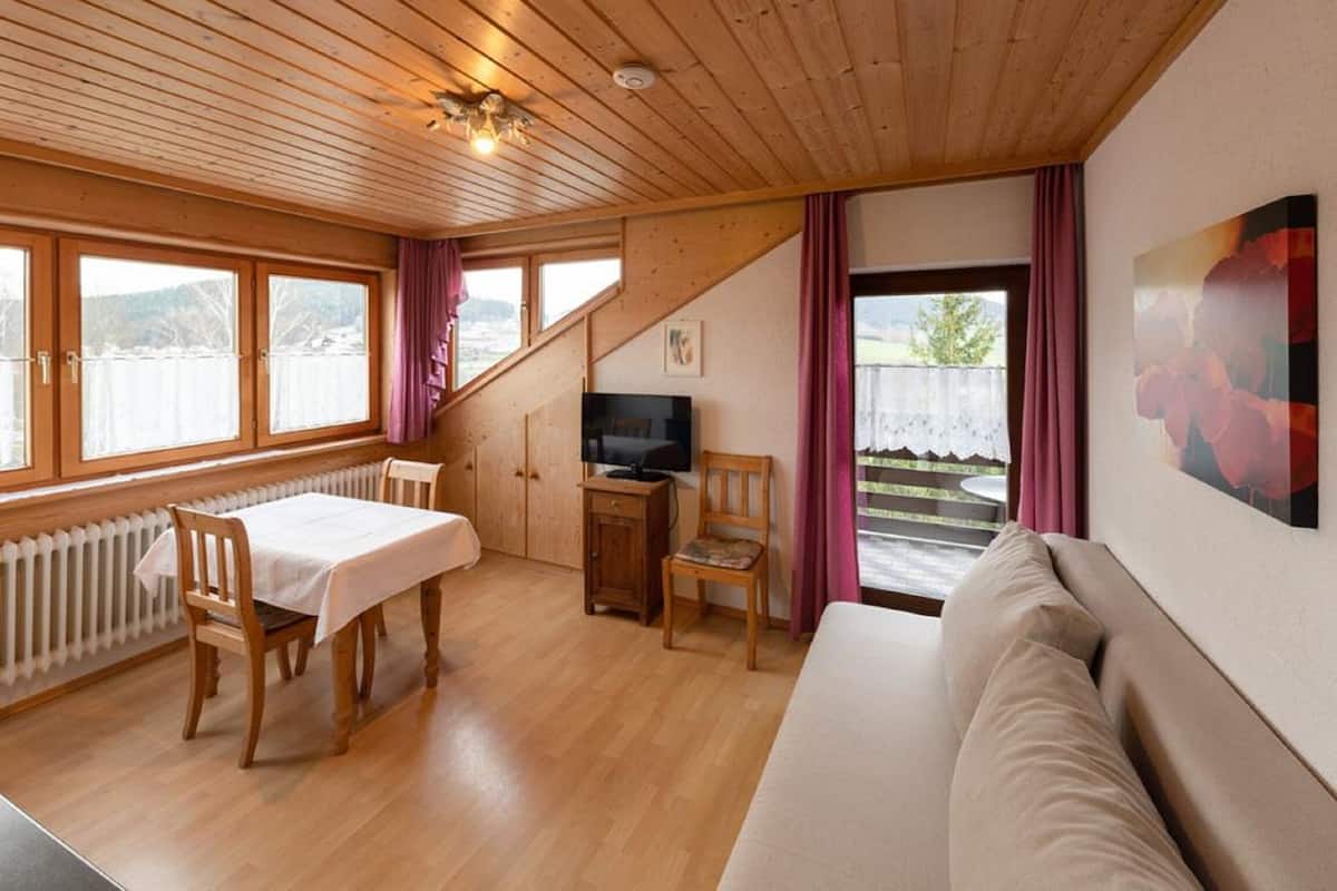 A cozy room with a wooden ceiling, a dining table with chairs, a sofa, a TV, and a view of a lake through large windows.