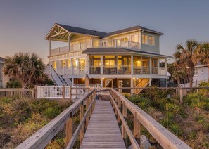 Une maison à deux étages située en bord de mer, avec une terrasse en bois menant à l’entrée.