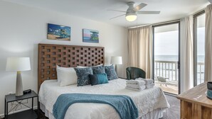 Une chambre d’hôtel avec un lit, deux tableaux, un ventilateur de plafond, une table de chevet, une chaise et une vue sur l’océan.