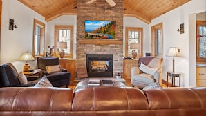A cozy living room with a stone fireplace, leather sofas, and a ceiling fan. - Grand Oaks B6- Mountain Condo, Gated Resort, Indoor/Outdoor Pools & Hot Tub (Boone)