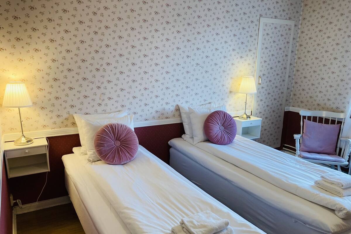 Une chambre d’hôtel avec deux lits simples, chacun doté de draps blancs et d’un oreiller violet. On y trouve des tables de chevet avec des lampes, une petite table en bois et une chaise avec un coussin. Les murs sont recouverts d’un papier peint à motifs floraux.