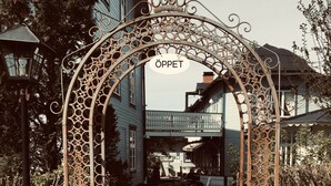 An ornate metal gate with the word "ÖPPET" above it, leading to a building with a porch and a large barrel.