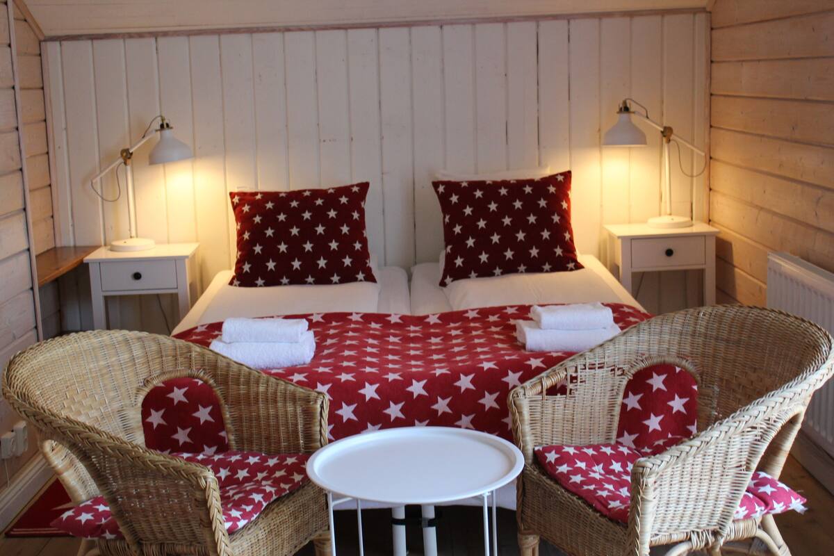 Un lit double avec des oreillers à motifs d’étoiles, deux tables de chevet avec des lampes, et deux fauteuils en osier avec des coussins à motifs d’étoiles.