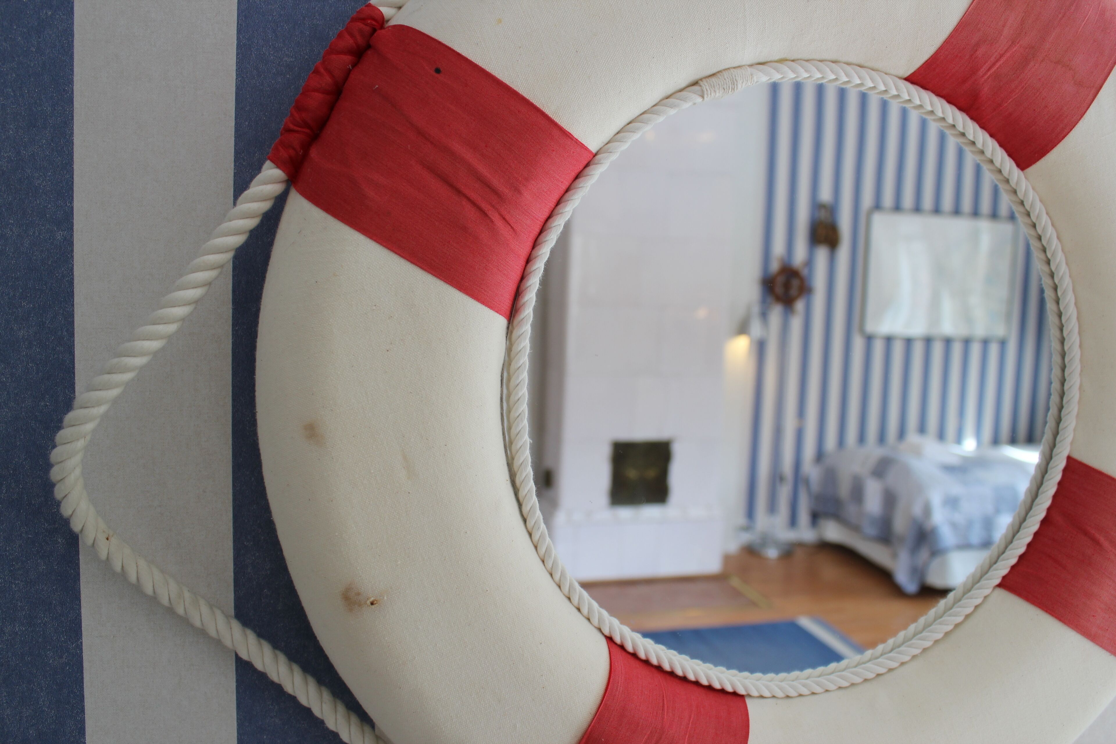 A circular mirror with a rope handle, reflecting a room with striped wallpaper and a bed.
