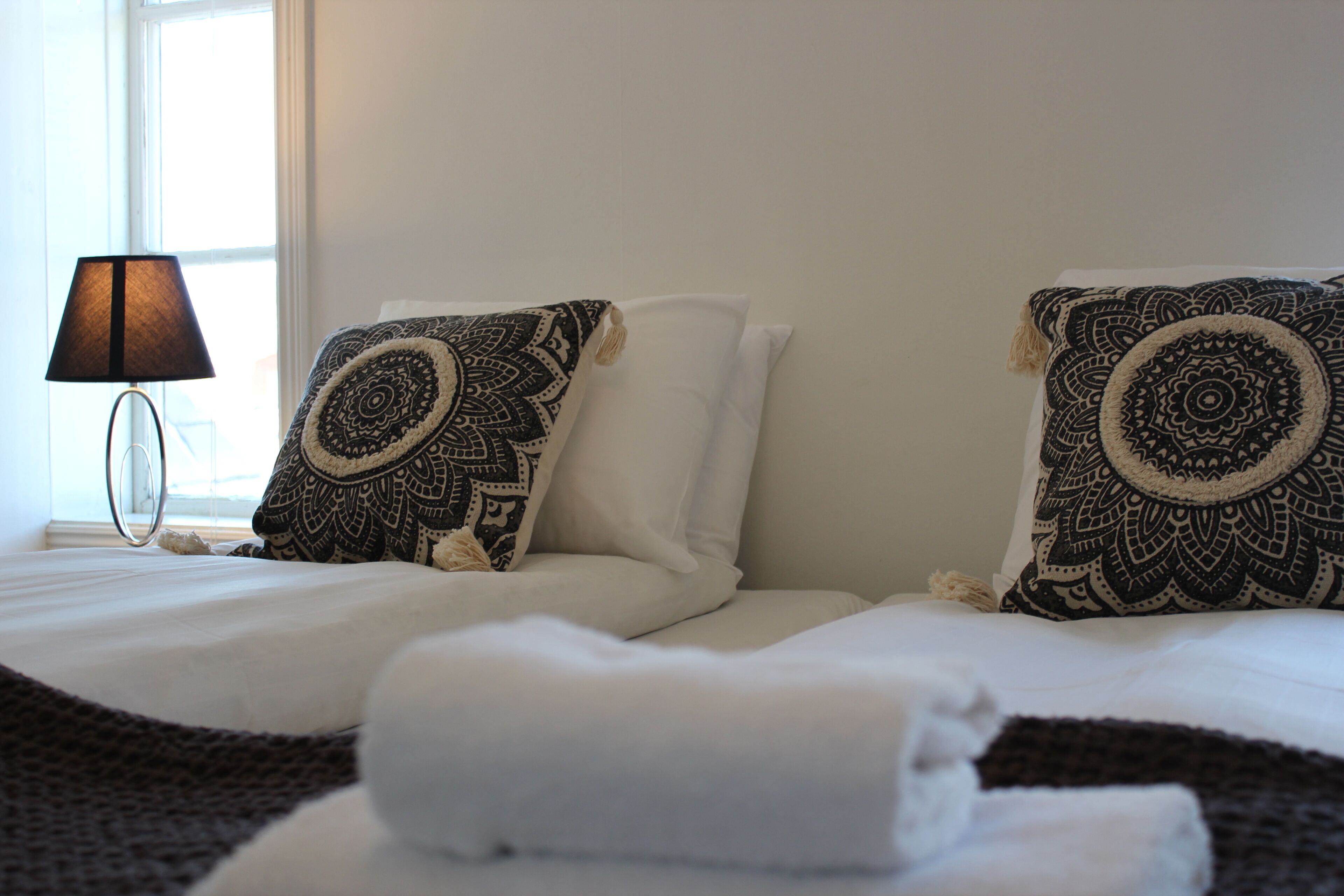 Una cama bien hecha con almohadones estampados y una toalla doblada, una lámpara de noche y una ventana con luz diurna.