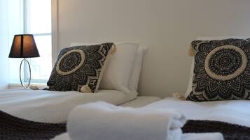 Una cama bien hecha con almohadones estampados y una toalla doblada, una lámpara de noche y una ventana con luz diurna.