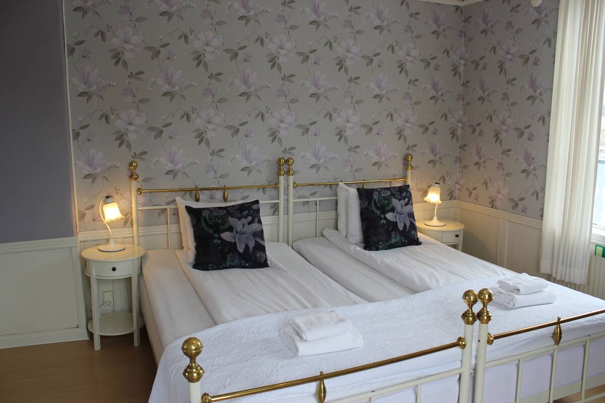 Un lit double avec du linge de lit blanc et des coussins à motifs floraux, deux tables de chevet avec des lampes et une fenêtre avec des rideaux.