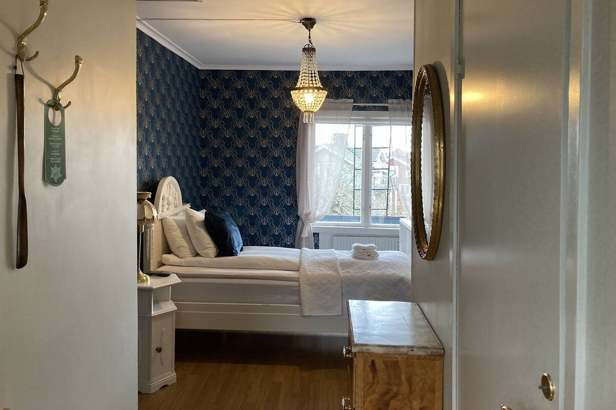 Une chambre à coucher comprenant un lit, une table de chevet, une commode, un lustre et une fenêtre avec des rideaux.