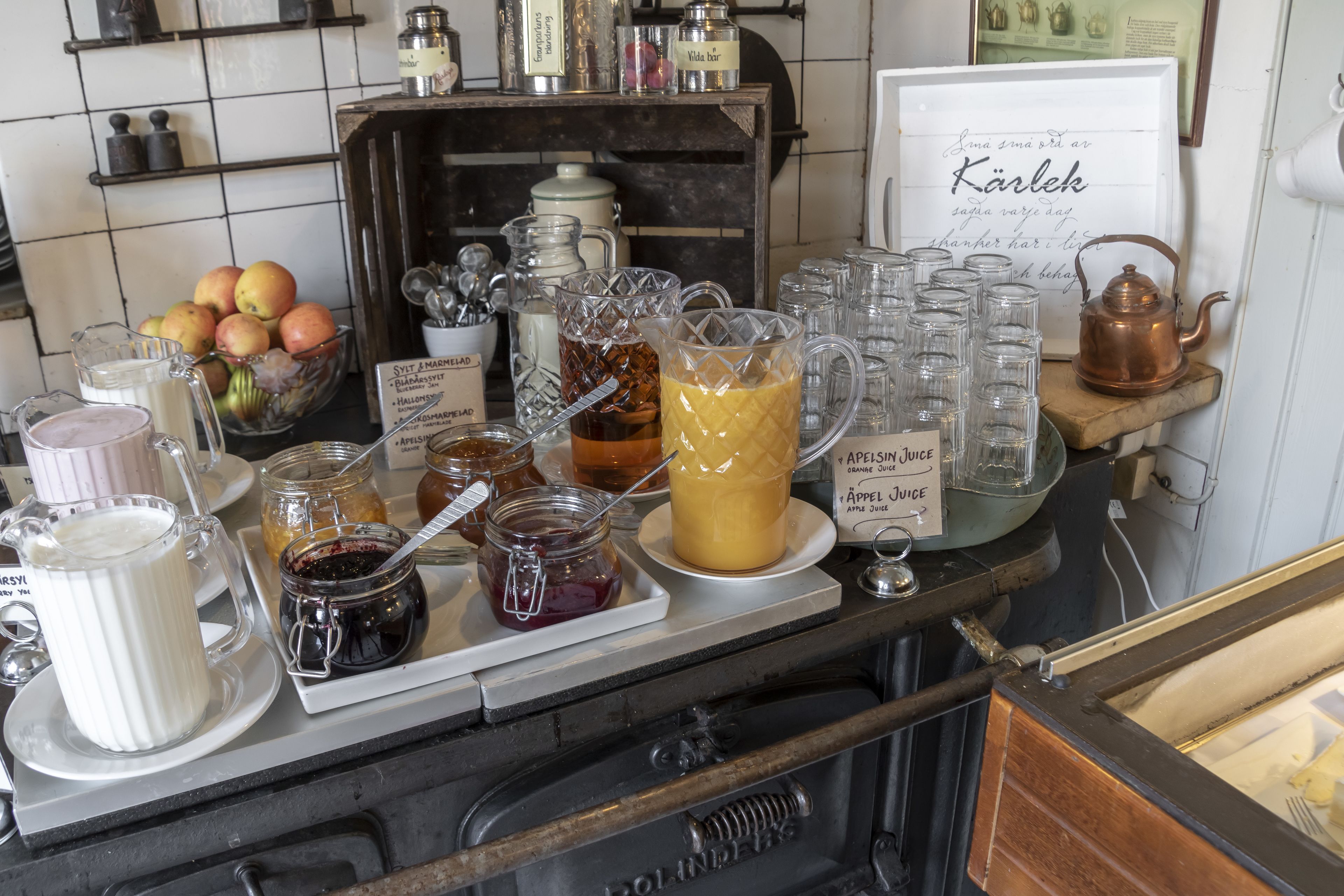 A breakfast bar with various food items and beverages, including jars of jam, a pitcher of milk, and a sign with a message in a foreign language.