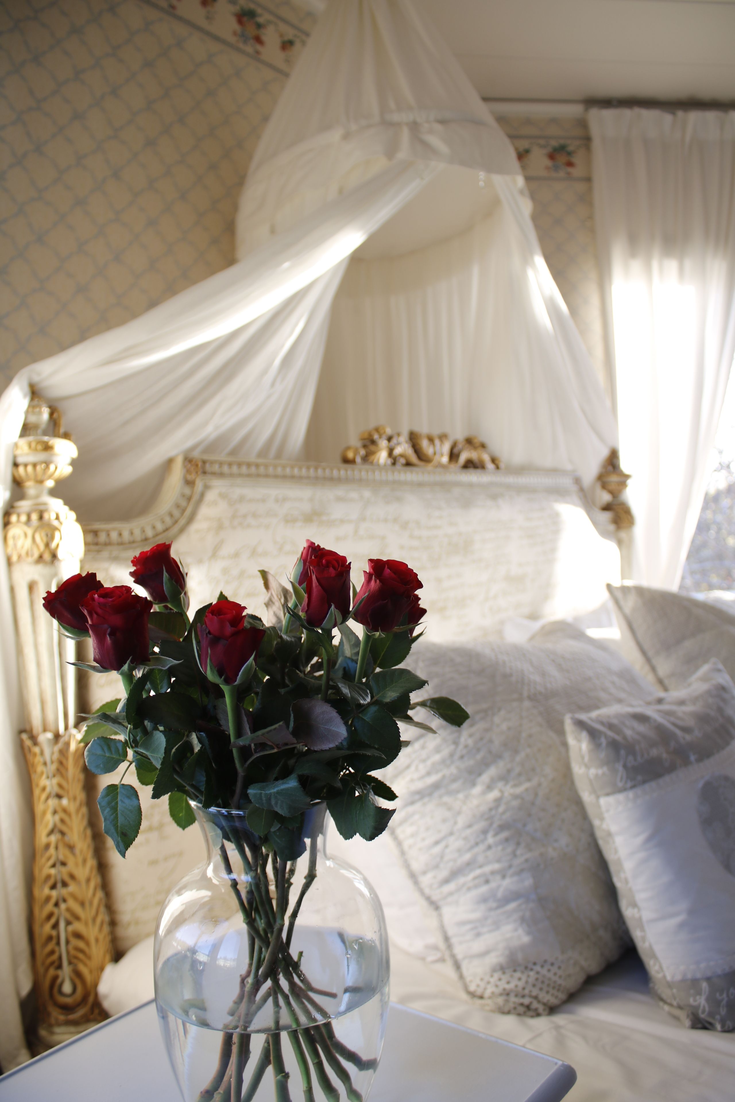 A bedroom with a canopy bed, a vase with red roses, and a decorative lamp.