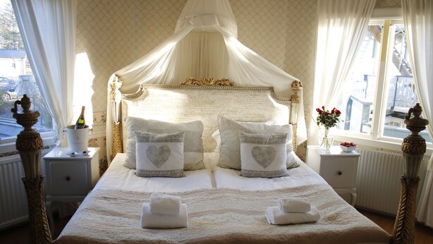 Een groot hemelbed met hartvormige kussens, een hemelbaldakijn en een nachtkastje met een fles champagne.