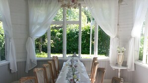Een lange eettafel gedekt voor een maaltijd, omringd door houten stoelen, met een kroonluchter en witte gordijnen.