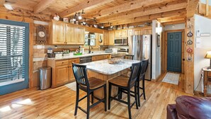 A spacious kitchen with wooden cabinets, a central island with chairs, stainless steel appliances, and a window with blinds. - A Portofino Cabin: Mountain Log Cabin, Hot Tub, Pet Friendly, Wood Fireplace (Seven Devils)
