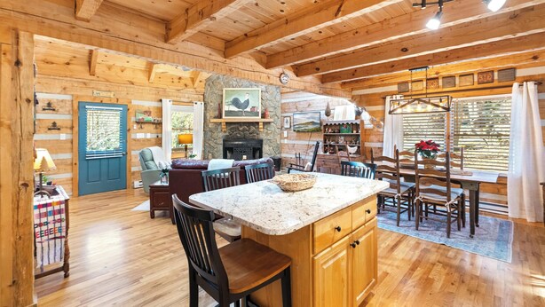 A cozy cabin interior with a stone fireplace, wooden dining area, and a large window with blinds. - A Portofino Cabin: Mountain Log Cabin, Hot Tub, Pet Friendly, Wood Fireplace (Seven Devils)