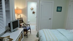 Makuuhuoneessa on sänky, jalustalla oleva televisio, tuoli, lamppu ja ovi, joka johtaa toiseen huoneeseen.