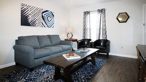 A living room with a blue sofa, a coffee table with books, a black leather chair, and a patterned rug. - Awesome Location 2 Bedroom 2.5 Bath / 511B (Lake Charles)