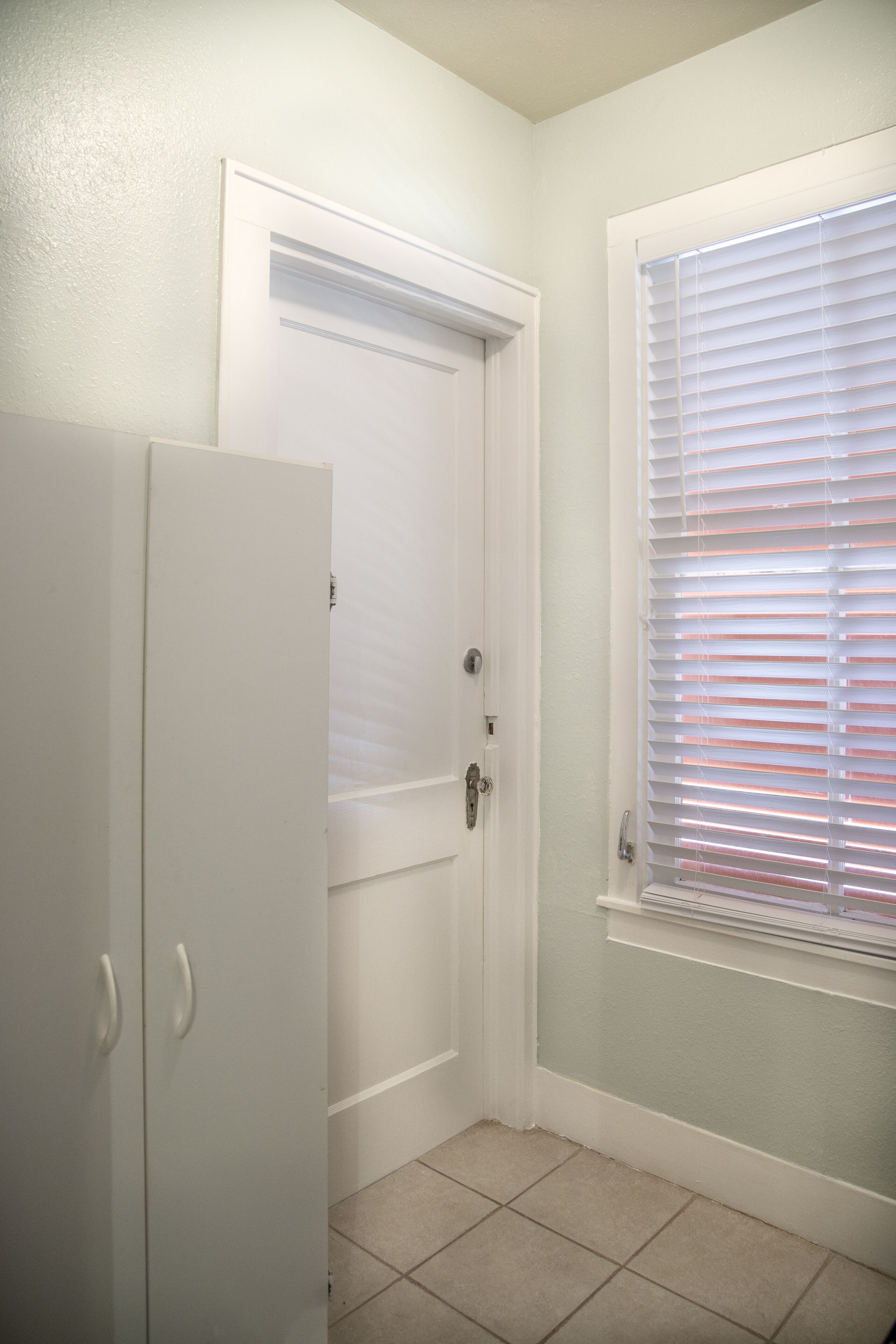 Una habitación pequeña y cerrada con una puerta blanca, una ventana con persianas y suelo de baldosas.